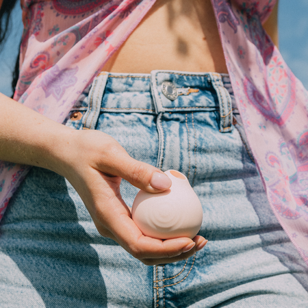 Woman's hand holding a HANX Cindy Pink Clitoral Stimulator, against a background of her bleach blue denim jeans and a crop top. It's a sunny day and the light is dappled on her skin.
