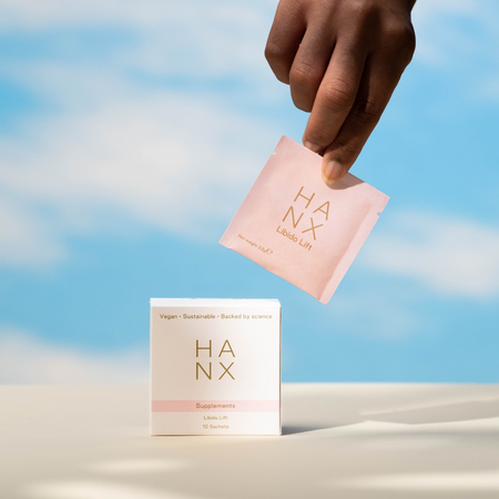 Brown skin toned hand holds a pink small sachet, which is branded with HANX logo in gold on the front, against a blue and white cloud sky background.