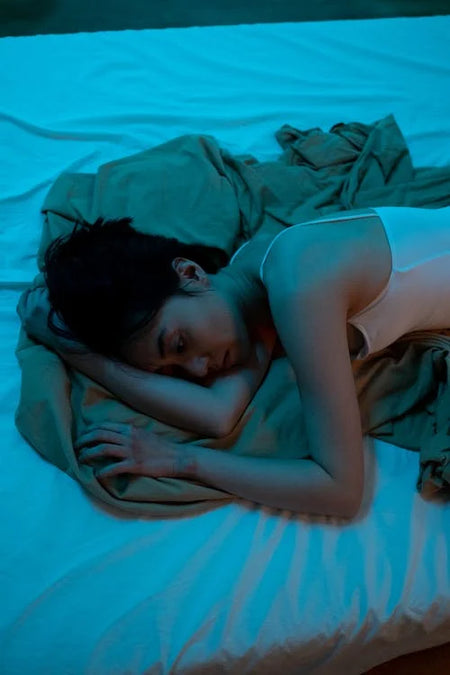 a woman looks upset and in pain laying curled up in bed with a blue hue 