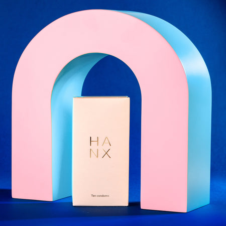 A gold and white 10 pack of HANX's vegan, sustainable, biodegradable condoms in the middle of a pink arch.