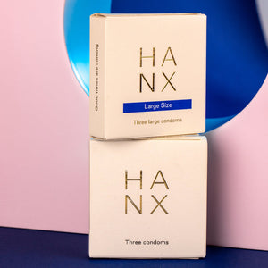 Two gold and white 3 pack of HANX's vegan, sustainable, biodegradable condoms stacked on-top of each other, one standard size and one large size.
