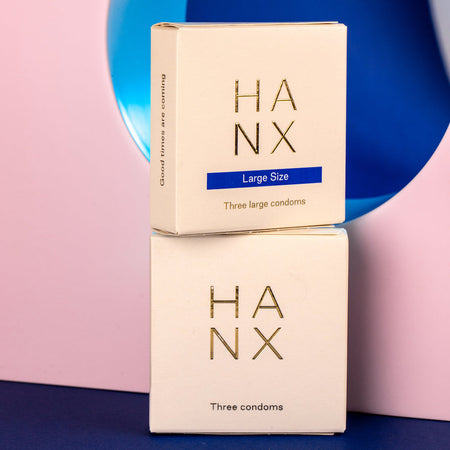 Two gold and white 3 pack of HANX's vegan, sustainable, biodegradable condoms stacked on-top of each other, one standard size and one large size.